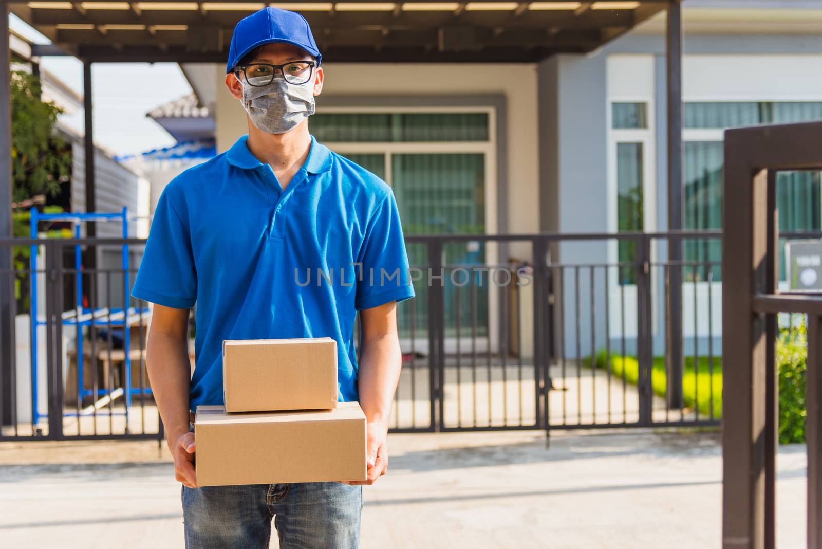 Asian Young delivery man courier online with box in blue cap and t-shirt uniform he protective face mask, service customer in front of the house under curfew quarantine pandemic coronavirus COVID-19