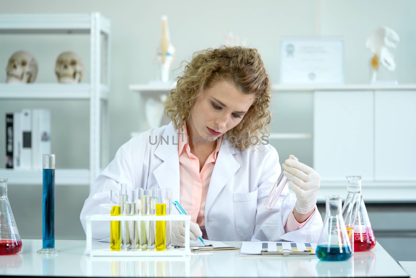 The young blonde scientist is writing down the result of new chemical research into the experiment chart. Working atmosphere in chemical laboratory. Test tubes filled with chemicals on the table.