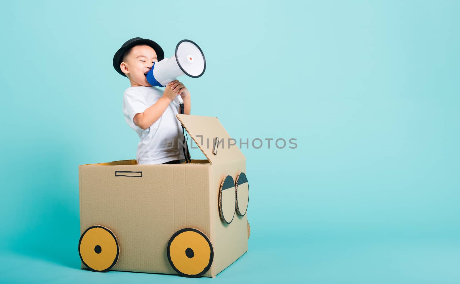 Happy Asian children boy smile in driving play car creative by a cardboard box imagination with megaphone, summer holiday travel concept, studio shot on blue background with copy space for text
