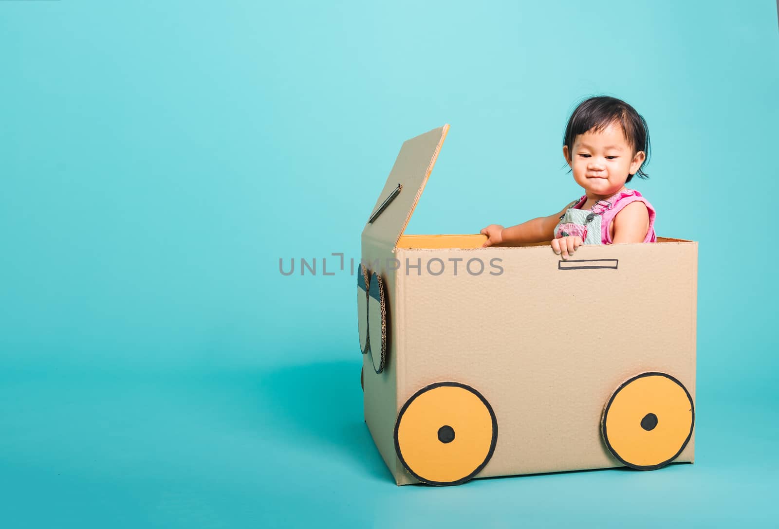 Happy Asian Baby girl smile in driving play car creative by a cardboard box imagination, summer holiday travel concept, studio shot on blue background with copy space for text