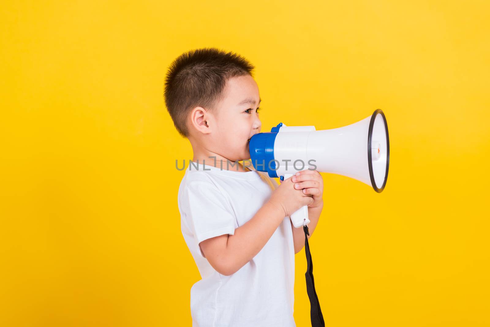 Asian Thai happy portrait cute little cheerful child boy holding and shouting or screaming through the megaphone her looking to side, studio shot isolated on yellow background with copy space