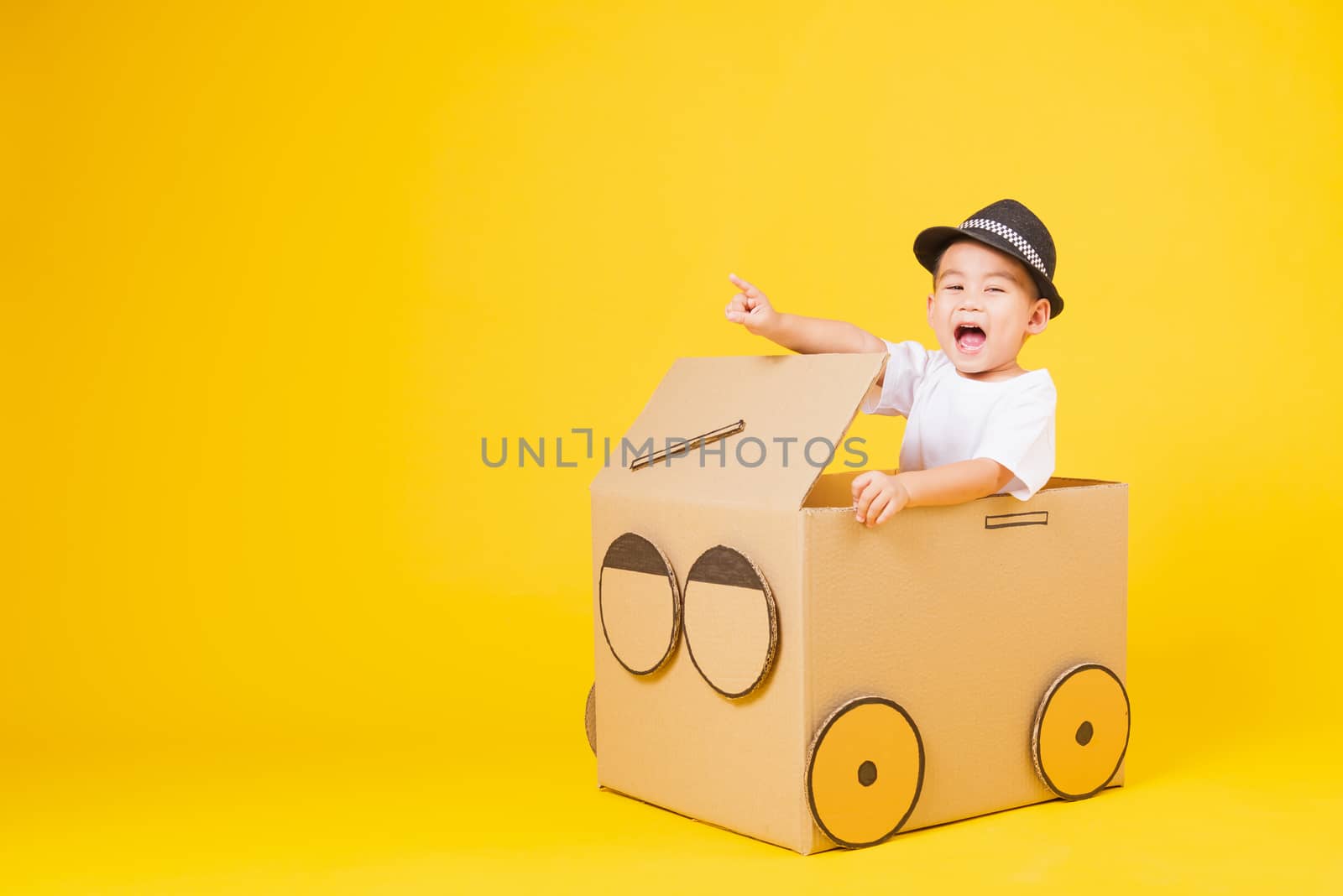 Portrait happy Asian cute little children boy smile so happy wearing white T-shirt driving car creative by cardboard and pointing finger, studio shot on yellow background with copy space