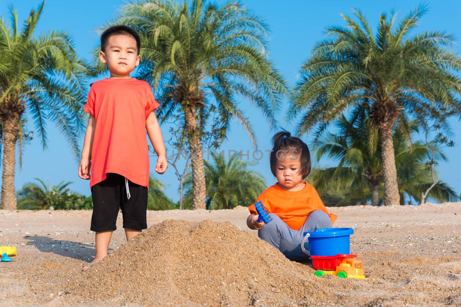 Asian Thai happy cute little cheerful Brother and sister two children funny digging play toy with sand at an outdoor tropical beach in summer day with copy space