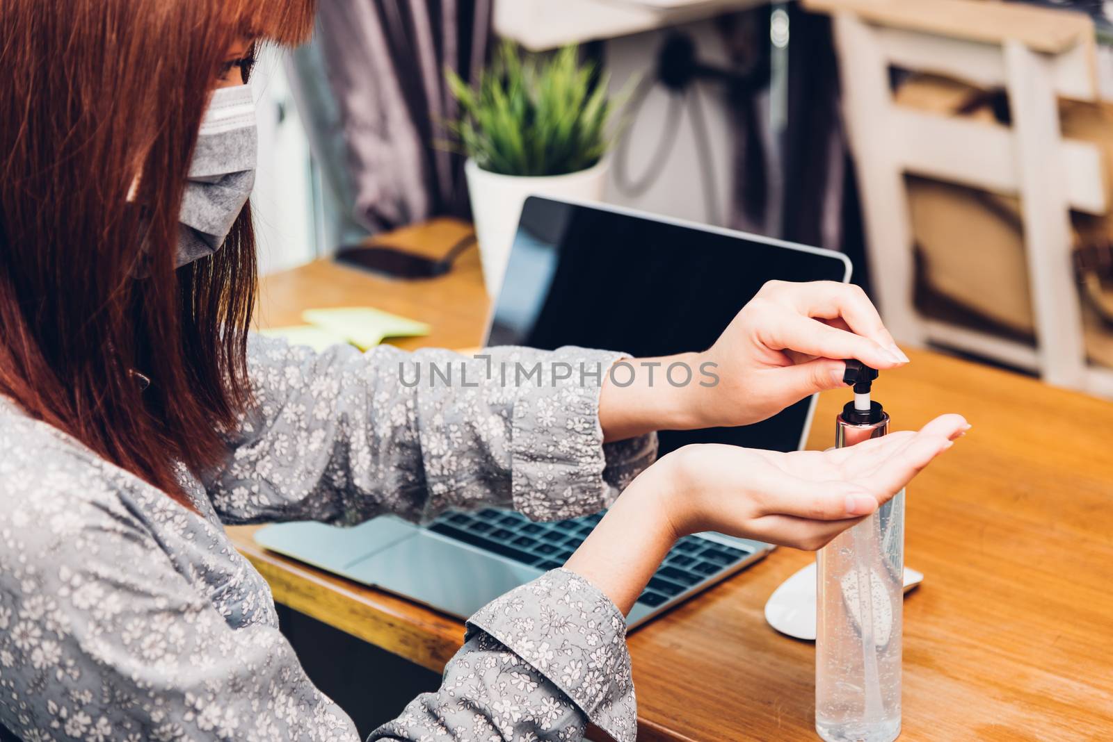 Asian Business young woman working from home office he quarantines disease coronavirus or COVID-19 wearing a protective mask and cleaning hands with sanitizer gel on front laptop computer