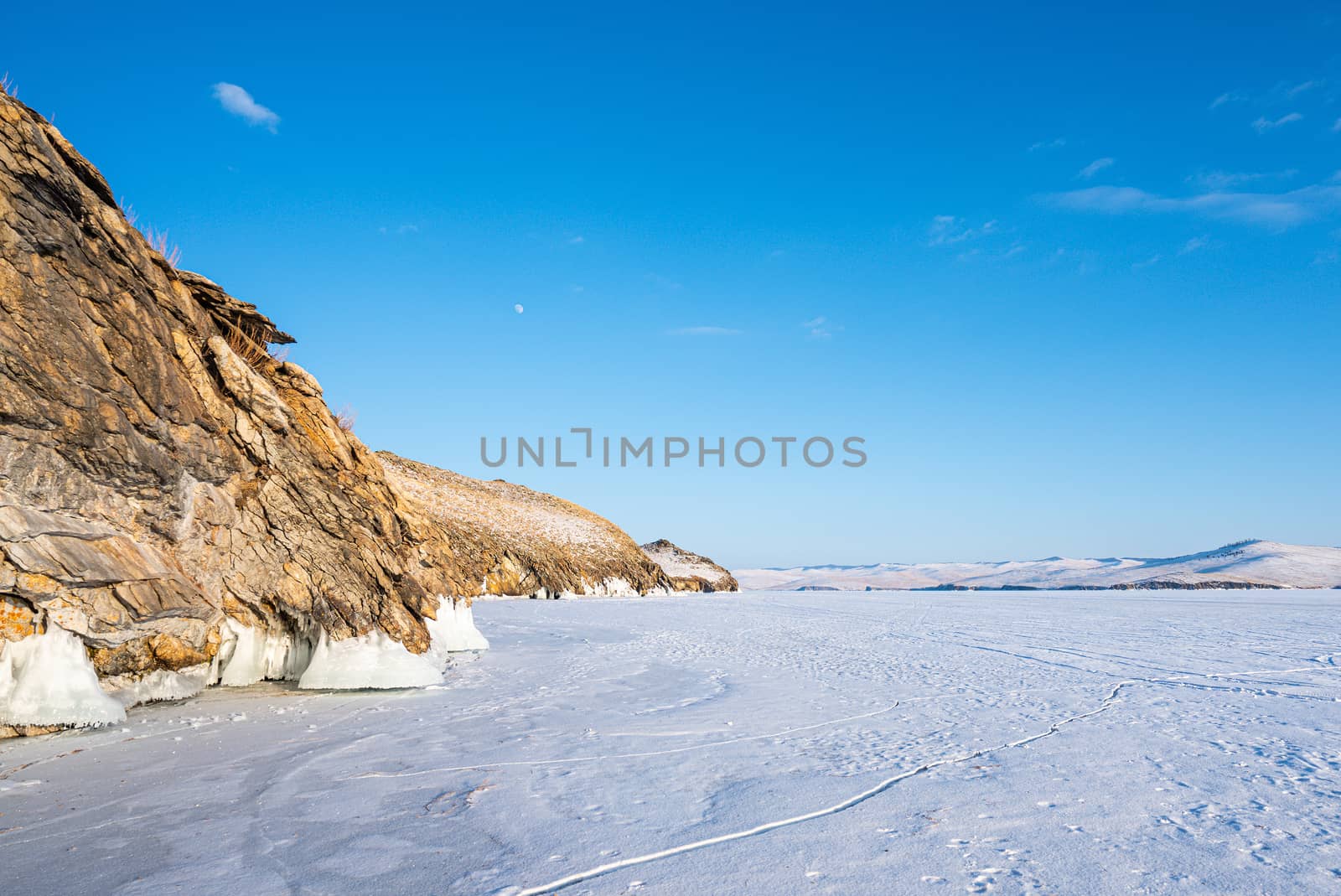 The rocky mountains are covered with snow. The blue color of the sky contrasts with the brown color of mountains and ground. Beautiful scenery of mountains, lake and skies. Lake Baikal, Russia.