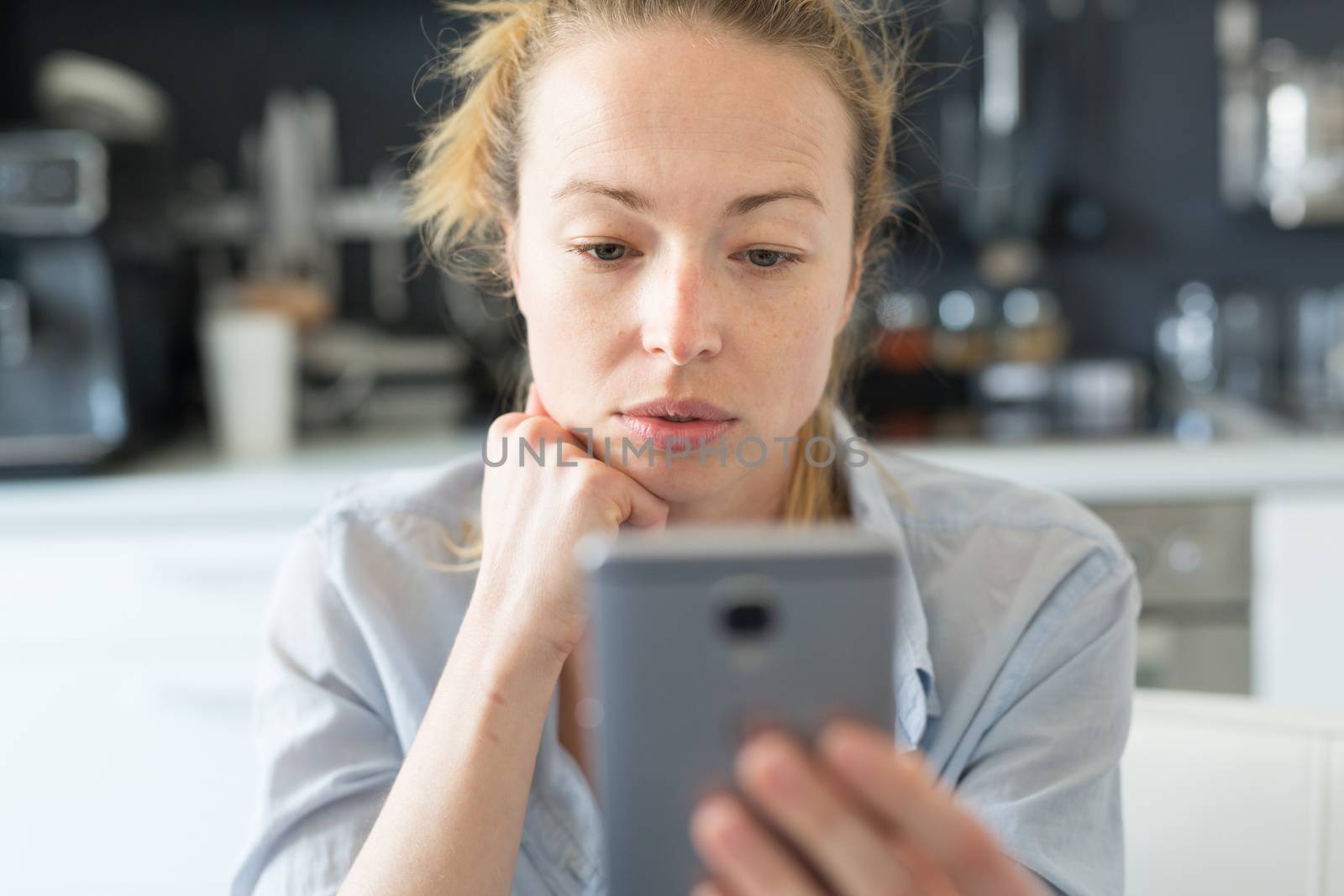 Young smiling cheerful woman indoors at home kitchen using social media apps on phone for video chatting and stying connected with her loved ones. Stay at home, social distancing lifestyle.
