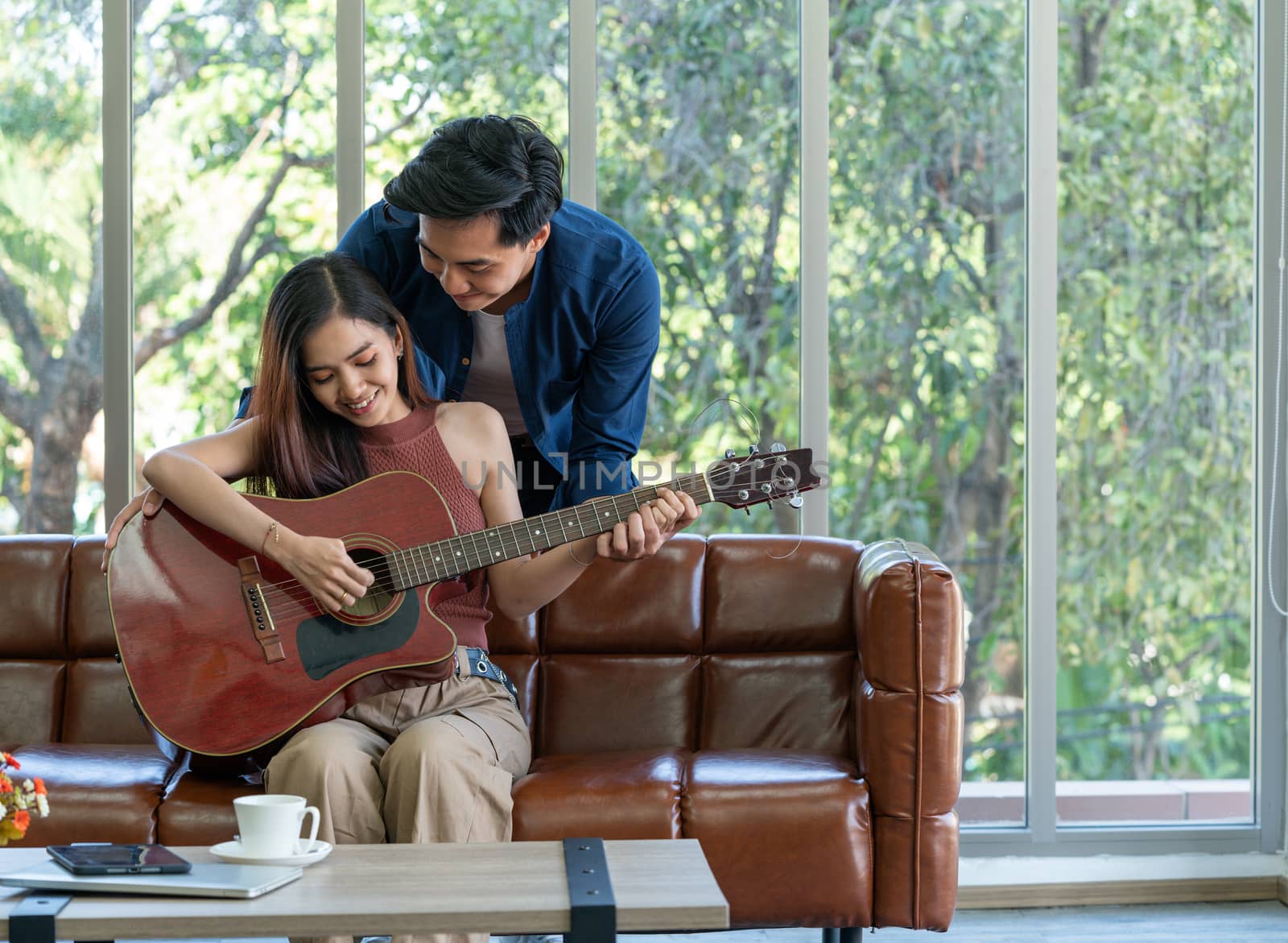 Young couples spend holidays in the living room. The young man wears comfortable clothes, teach his girlfriend to play a guitar. Asian woman enjoy playing music instrument.