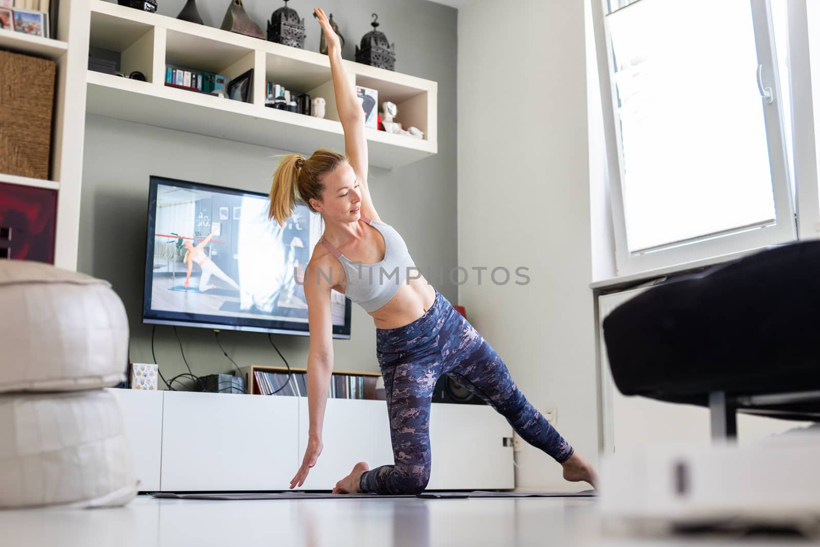 Attractive sporty woman working out at home, doing pilates exercise in front of television in her living room. Social distancing. Stay healthy and stay at home during corona virus pandemic.