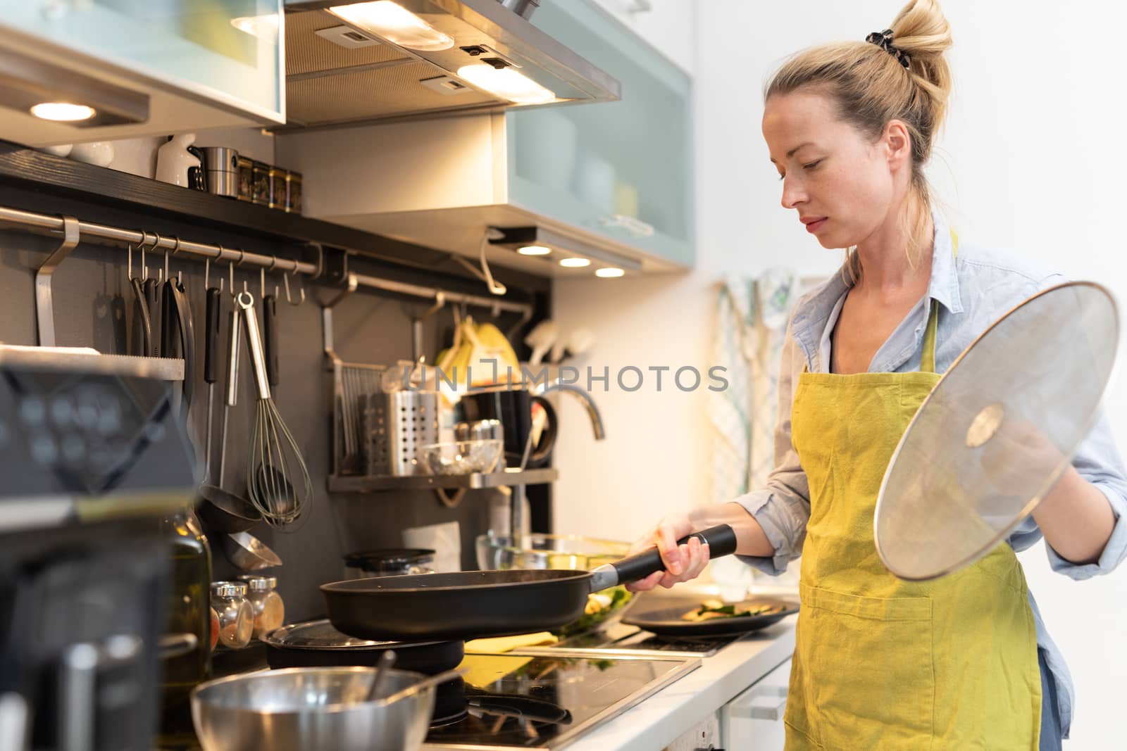 Stay at home housewife woman cooking in kitchen, stir frying dish in a saucepan, preparing food for family dinner. Blonde caucasian model in her 30is, real cooking not looking in camera.
