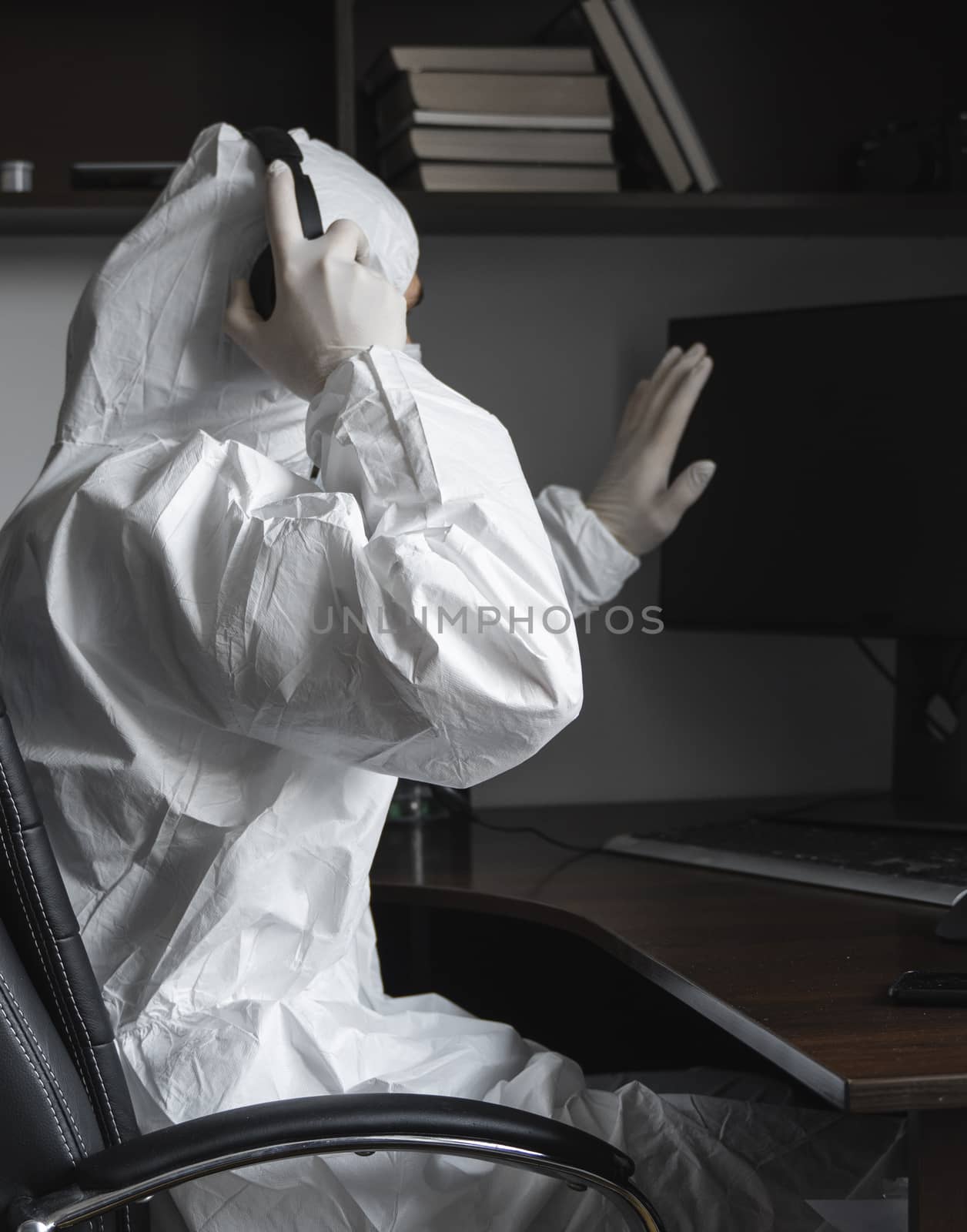 Man in medical mask and protective suit, gloves sits at home and works with pc at the table during quarantine and show hello to his business partner or friends. Remote work in a pandemic covid