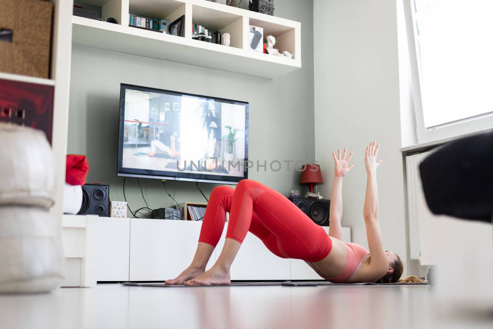 Attractive sporty woman working out at home, doing pilates exercise in front of television in small studio appartment. Social distancing. Stay healthy and stay at home during corona virus pandemic.