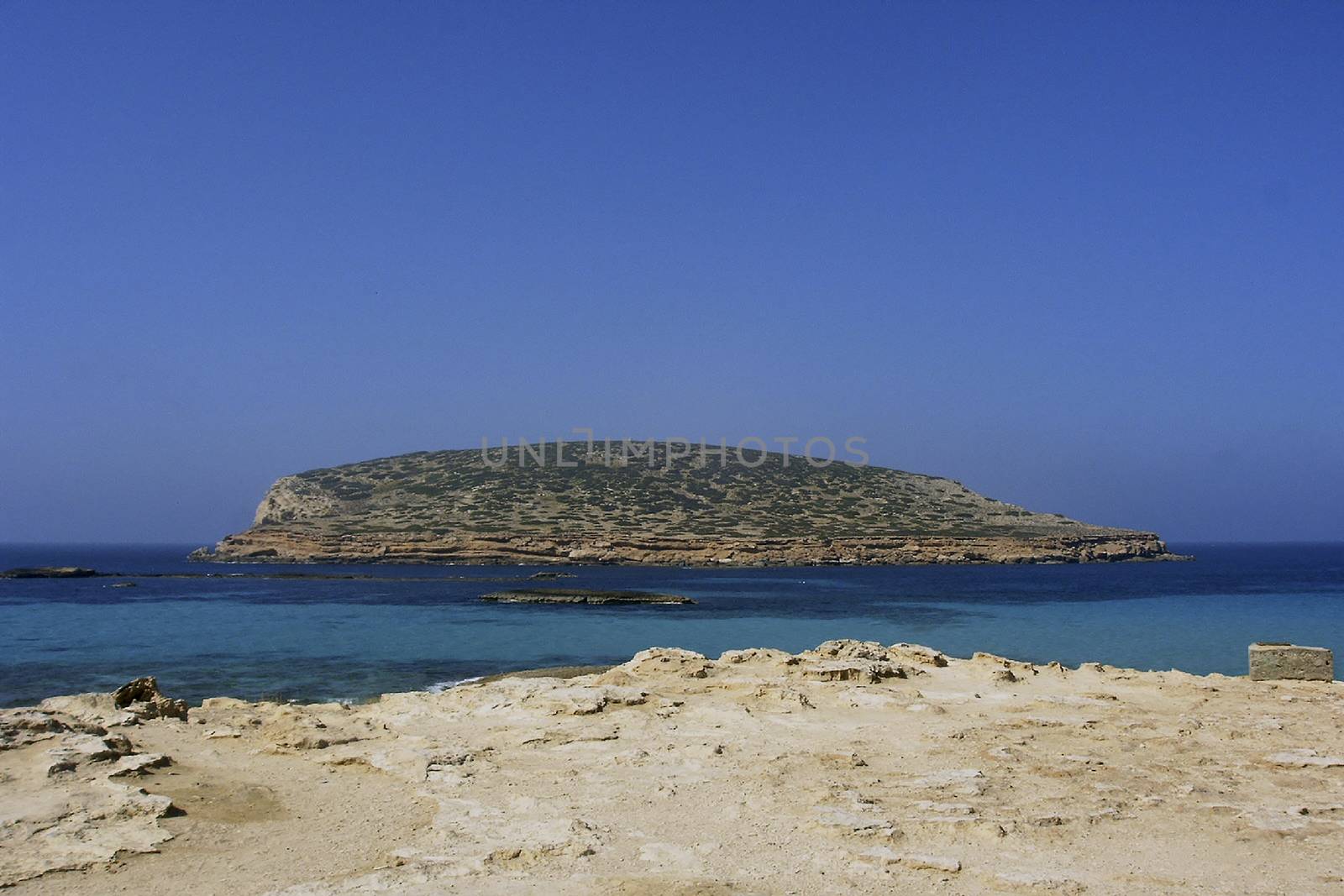 Deserted beach with turquoise waters, bright day, Mountains in the background and bright blue day, with a large rock in the middle