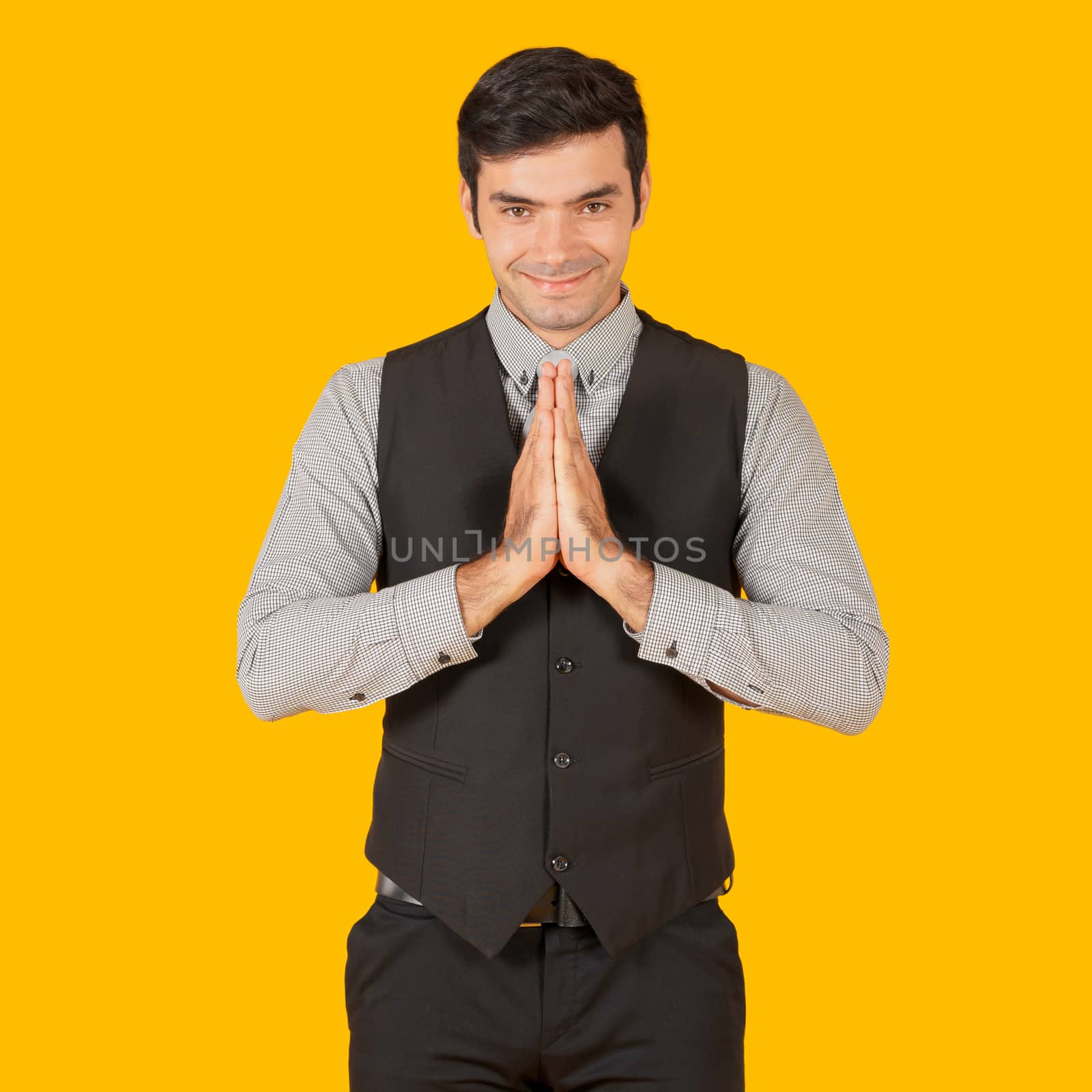 Young businessman in a gray shirt and black vest press the hands together at the chest in sign of respect, smiling and bowing a little. Concept of business relationships. Portrait on yellow background