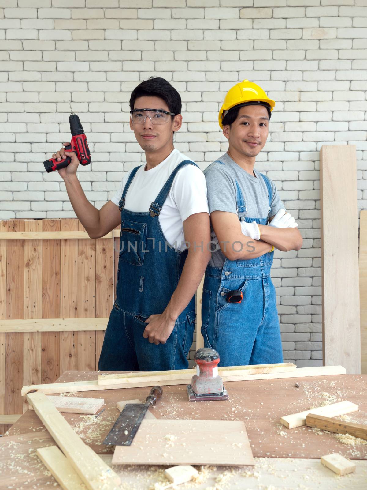 Two Asian carpentry poses confidently before beginning the job of receiving orders at the wood working place. Morning work atmosphere in the workshop room. A desk full of hand tools and wood piles.