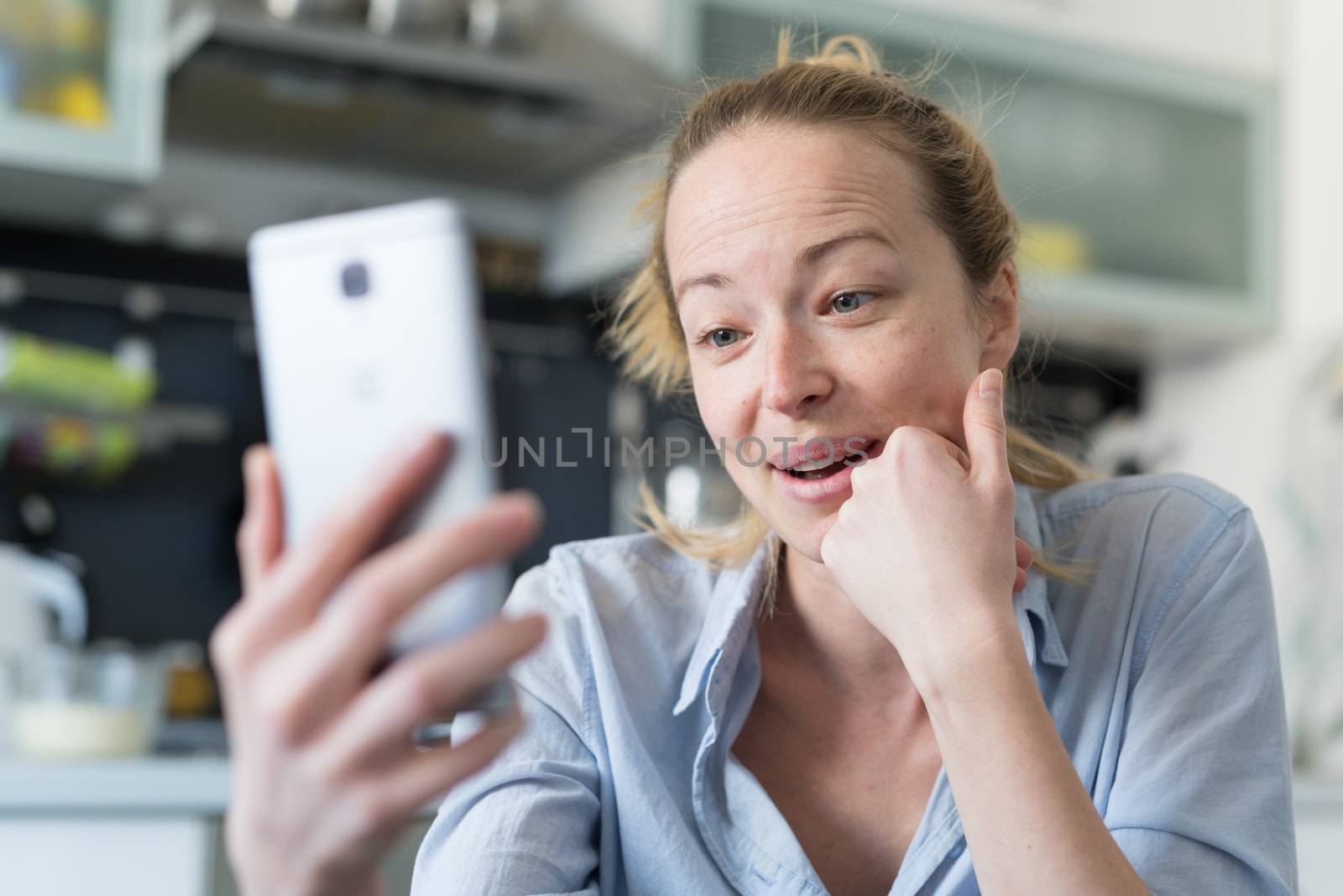 Young smiling cheerful woman indoors at home kitchen using social media apps on phone for video chatting and stying connected with her loved ones. Stay at home, social distancing lifestyle.