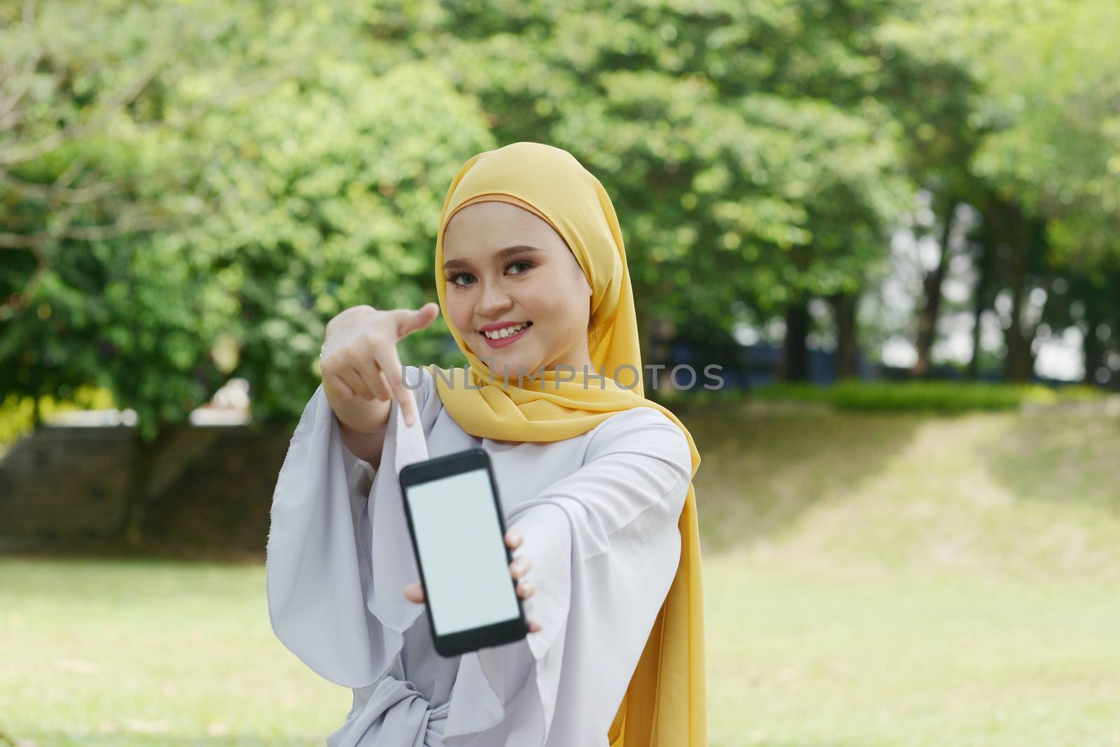 Portrait of cheerful Muslim girl hand pointing at smartphone, smiling at outdoor.