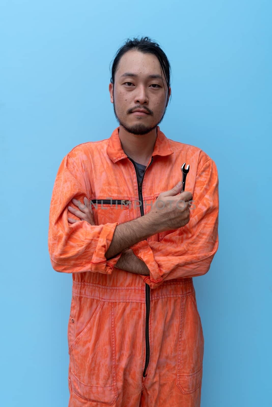 The chief mechanic in an orange uniform held a small wrench. Standing with his arms crossed on the blue screen. Portrait with studio light.