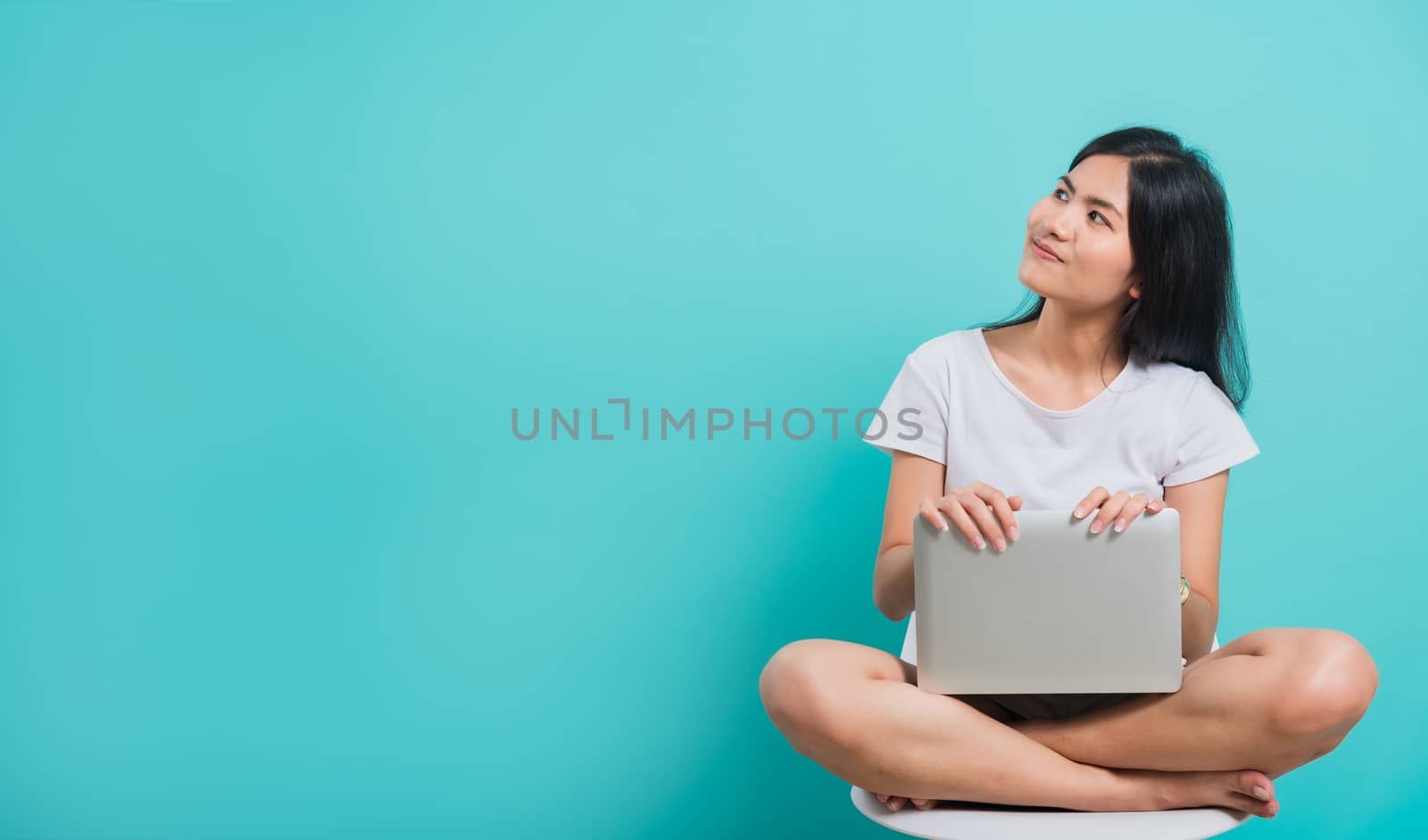 Portrait happy Asian beautiful young woman smile with teeth sitting on chair wear white t-shirt, She holding and using a laptop computer, studio shot on blue background with copy space for text