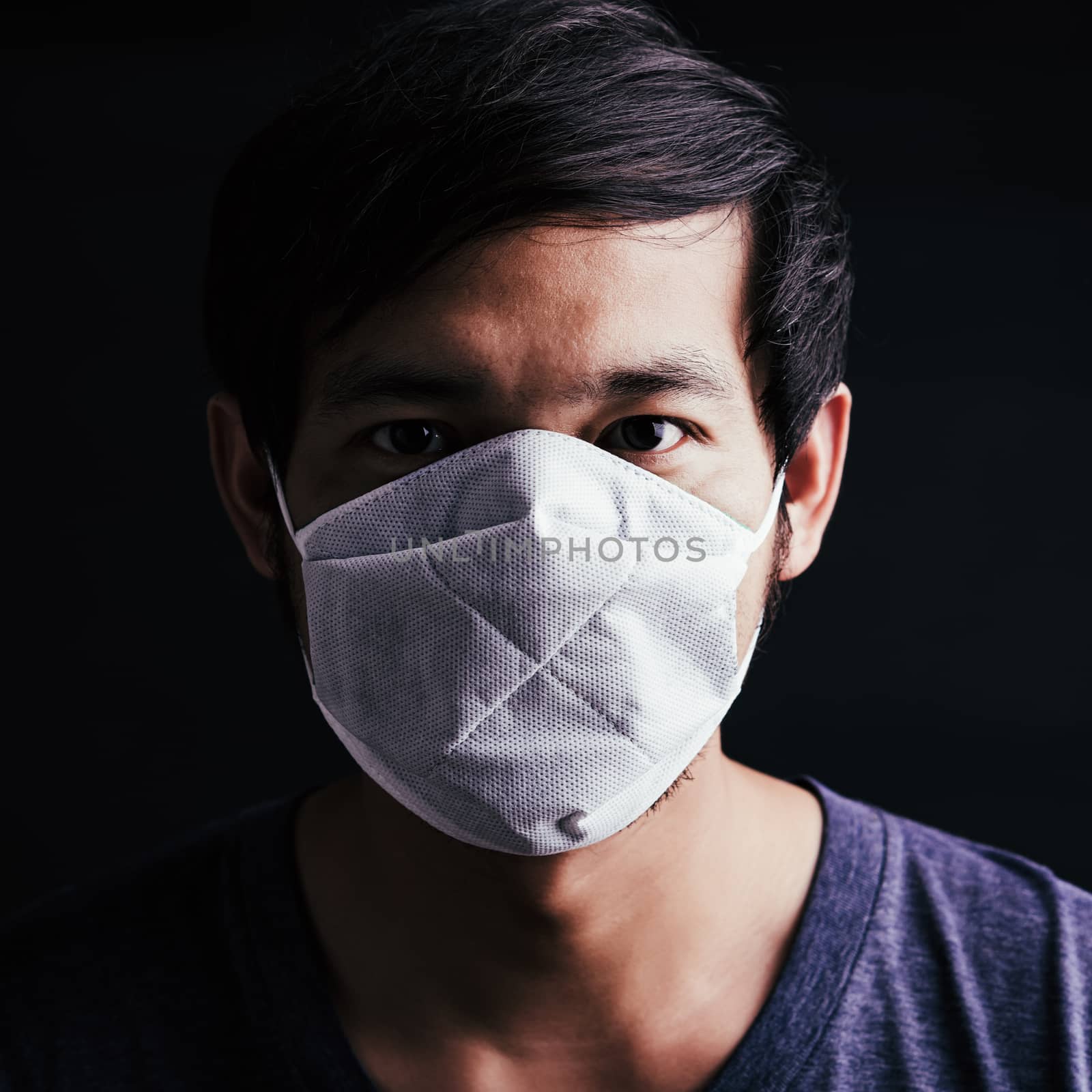 Closeup Asian young man wearing protective face mask with fear in the eye, prevent germs or disease hygiene prevention COVID-19 virus or coronavirus protection concept, dark on black background