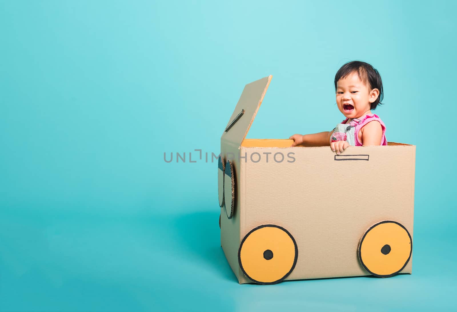 Happy Asian Baby girl smile in driving play car creative by a cardboard box imagination, summer holiday travel concept, studio shot on blue background with copy space for text