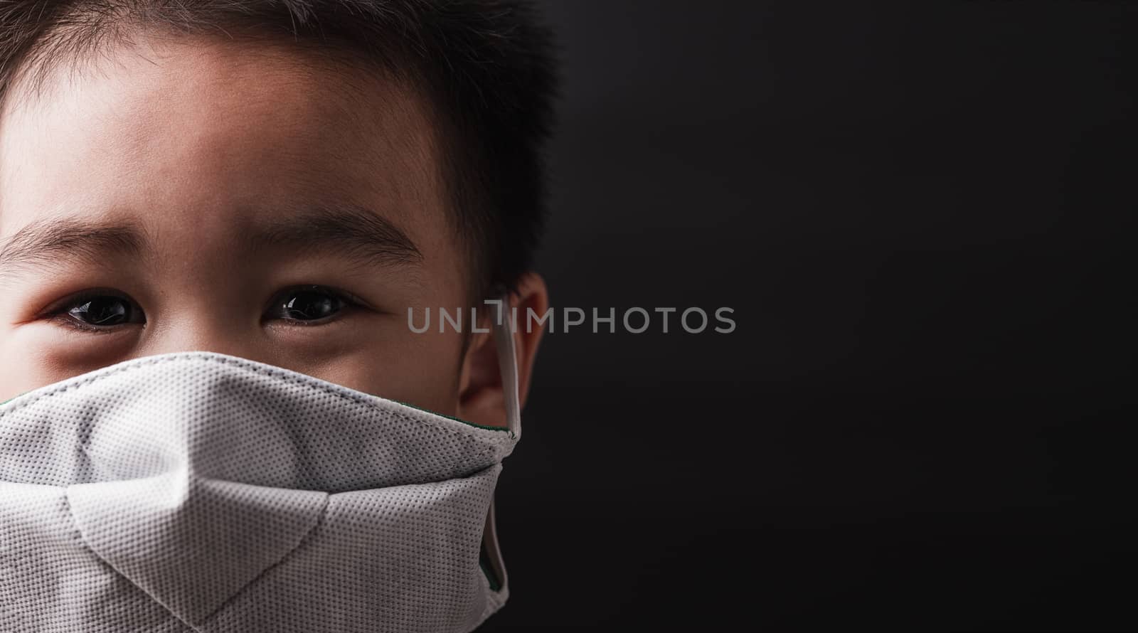 Closeup Asian little child wearing protective face mask with fear in the eye, prevent germs or disease hygiene prevention COVID-19 virus or coronavirus protection concept, dark on black background