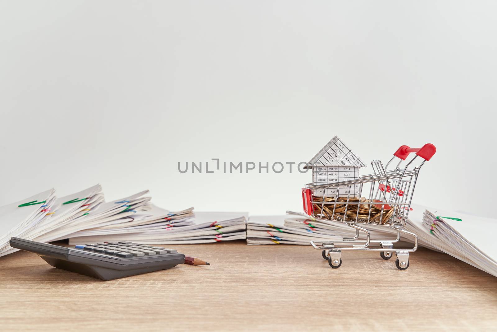 House on gold coins in shopping cart and calculator with pencil on wooden table have pile of paperwork of report of sale and receipt with colorful paperclip as background and copy space.