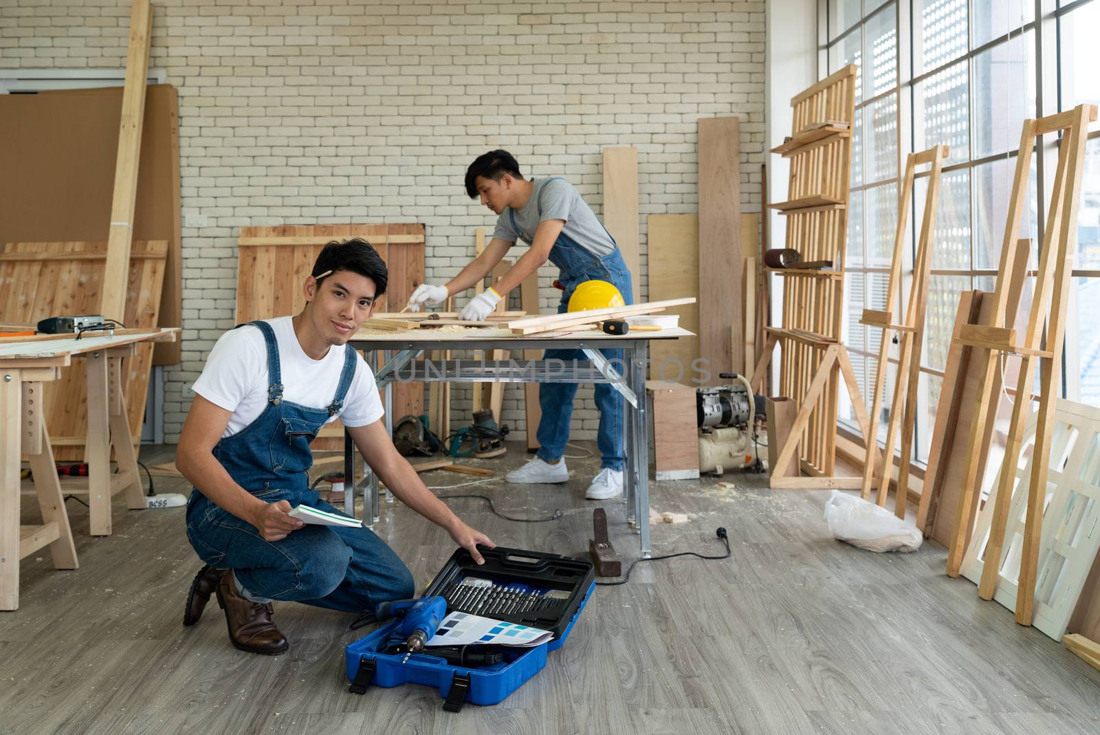 Young asian carpenter sat with the equipment confidently in his workshop. Another man using a pencil to draw drawings on a piece of wood. 