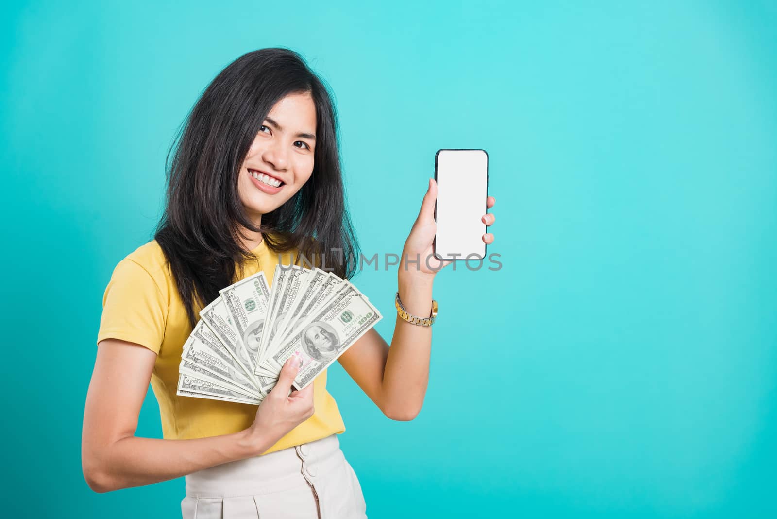 Asian happy portrait beautiful young woman standing wear t-shirt smile holding money fan banknotes 100 dollar bills using mobile phone and looking to camera on blue background with copy space for text