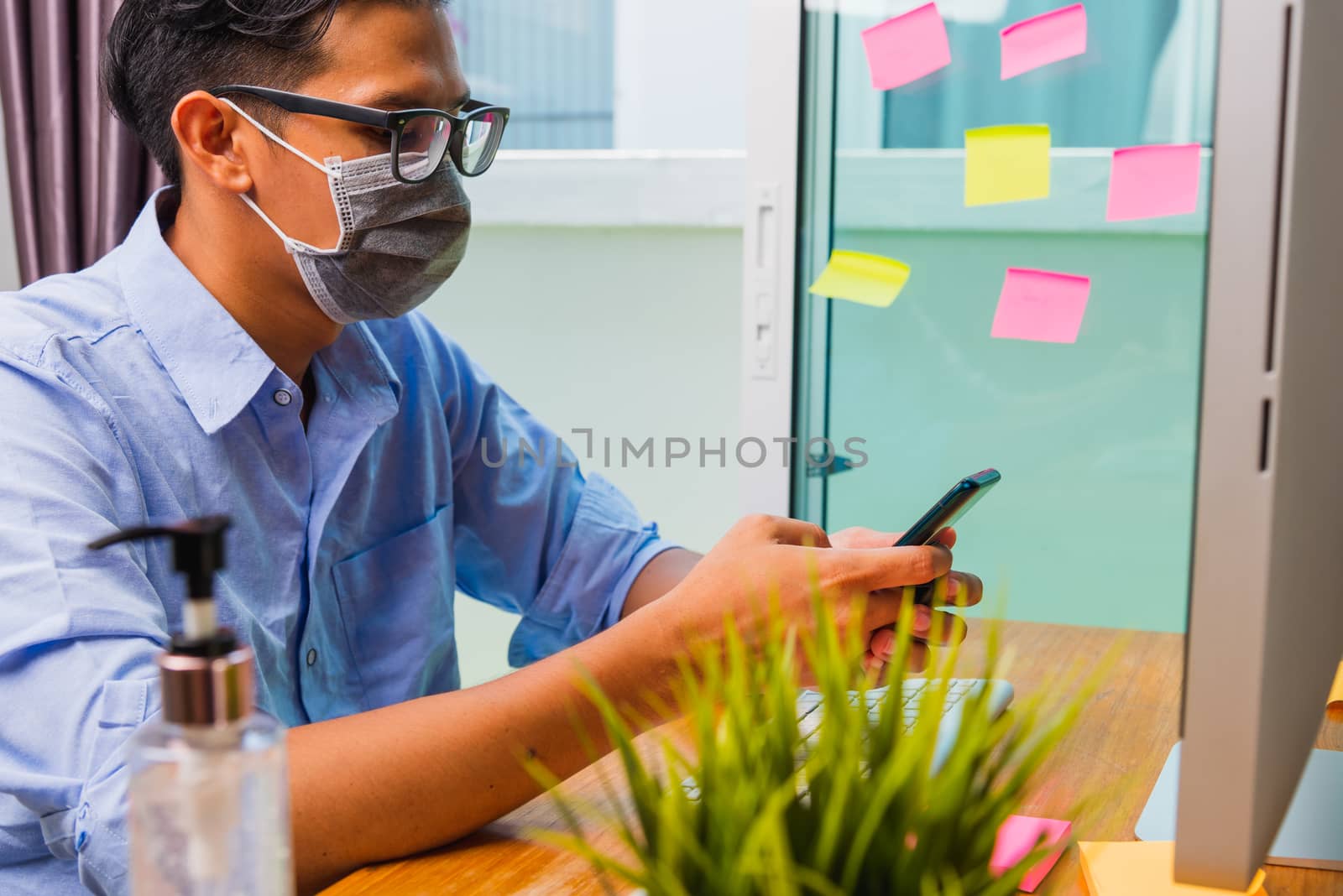 Asian business young man wearing a protective mask with disinfectant gel beside, work from home office with PC computer he using mobile smart phone, quarantines disease coronavirus or COVID-19