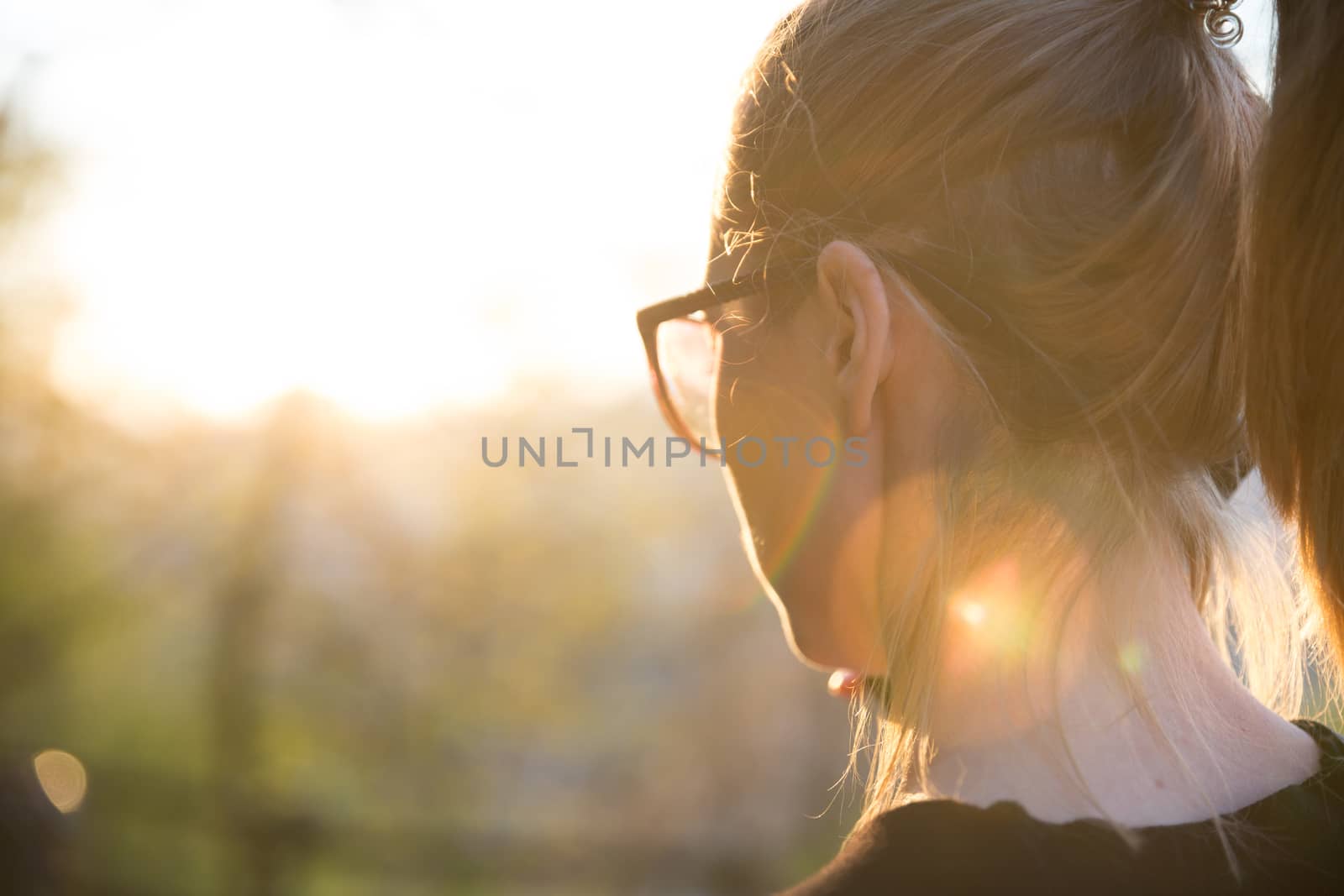 Backlit rear view of young woman talking on cell phone outdoors in park at sunset. Girl holding mobile phone, using digital device, looking at setting sun. Copyspace.