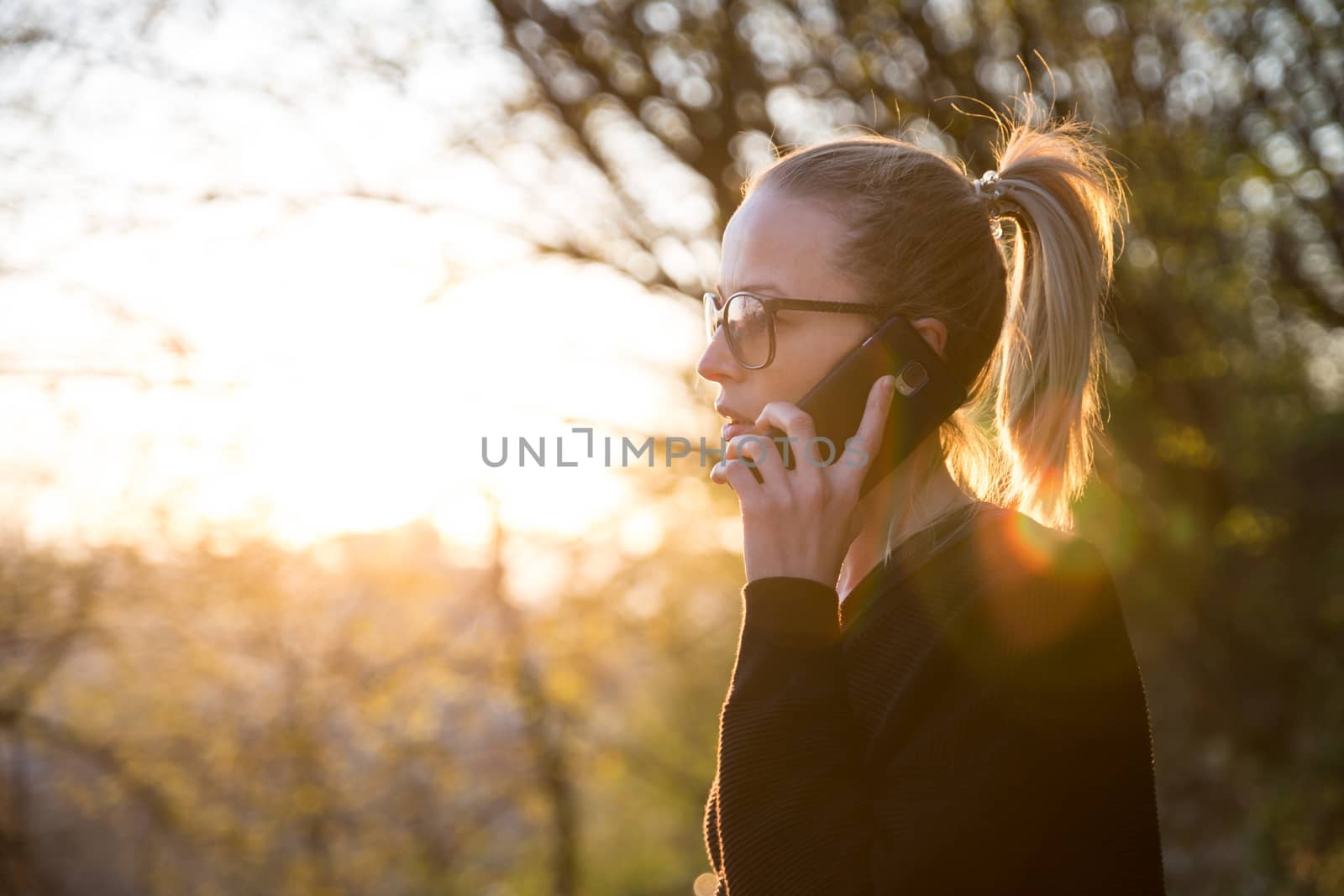Backlit rear view of young woman talking on cell phone outdoors in park at sunset. Girl holding mobile phone, using digital device, looking at setting sun. Copyspace.