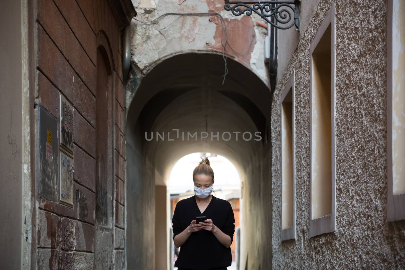 COVID-19 pandemic coronavirus. Casual caucasian woman at medieval city street using mobile phone, wearing protective face mask against spreading of coronavirus and disease transmission.