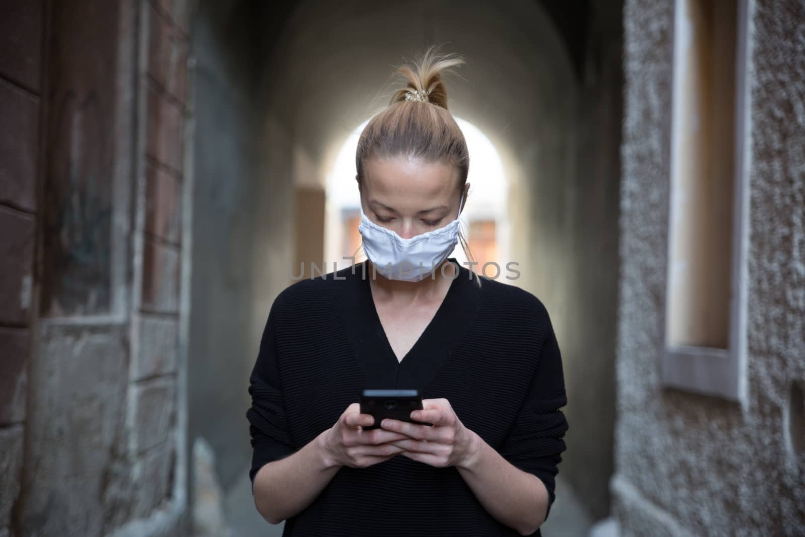 COVID-19 pandemic coronavirus. Casual caucasian woman at medieval city street using mobile phone, wearing protective face mask against spreading of coronavirus and disease transmission.