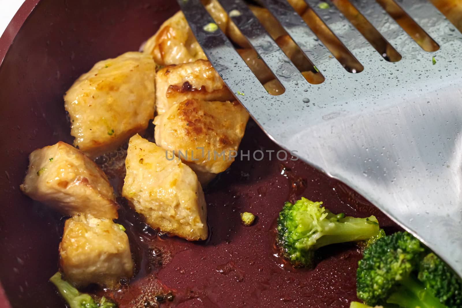 In a frying pan, vegetarian simulated chicken and fresh broccoli are being cooked. A close-up view shows the detail in the food, bubbling oils on the pan, and the metal spatula flipper leaning across the top.