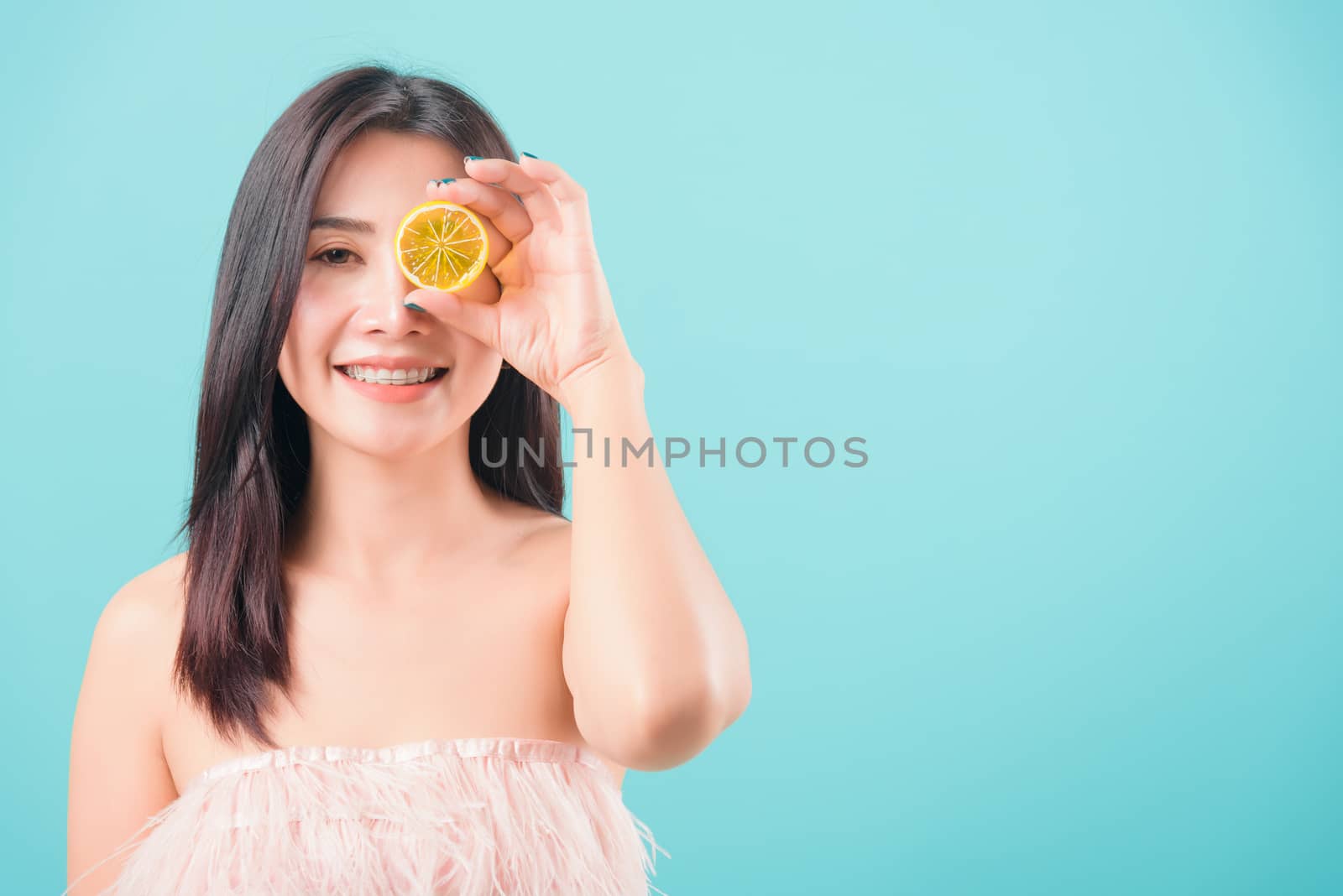 Asian happy portrait beautiful young woman standing smile holding a piece of orange in front of her eye on blue background with copy space for text, healthy lifestyle concept