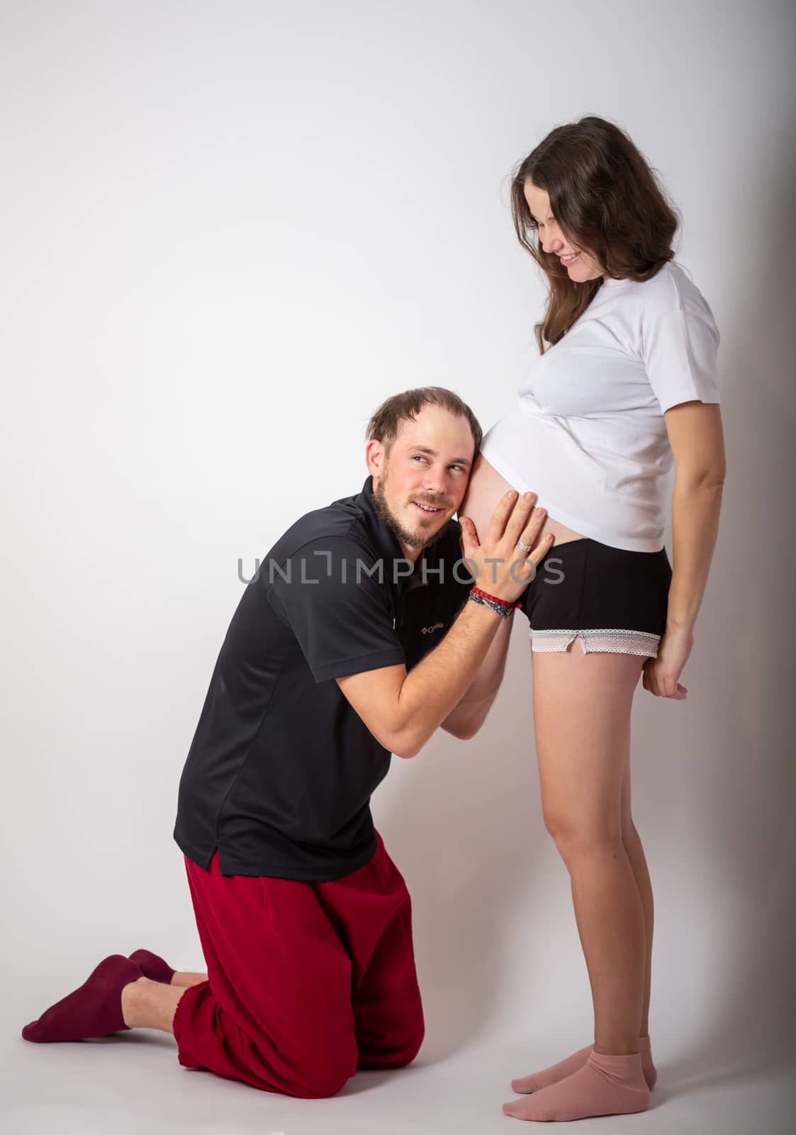 Handsome man is listening to his beautiful pregnant wife's tummy and smiling.