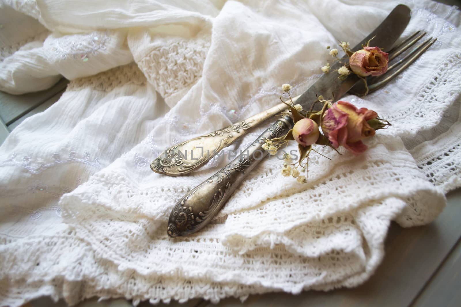 Light pastel colored tableware set: vintage silverware on napkin and delicate pink dry roses on rustic shabby wooden table