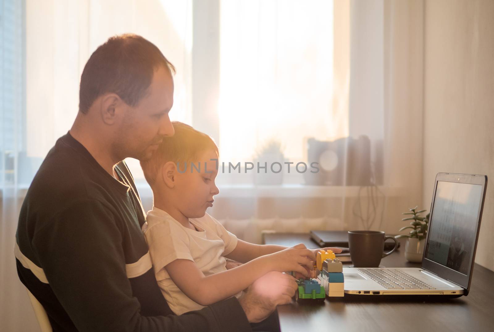 Four year old boy play and father at the table with notebook. Home working at coronavirus quarantine isolation period. Working among children, working remote concept. Sunset light