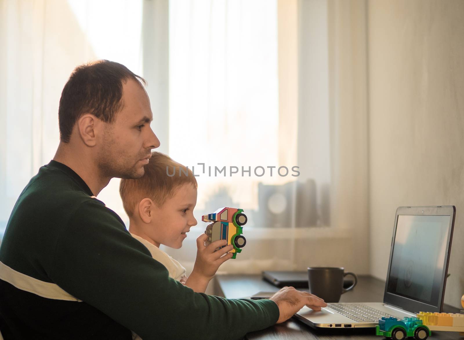 Four year old boy play and father at the table with notebook. Home working at coronavirus quarantine isolation period. Working among children, working remote concept. Sunset light