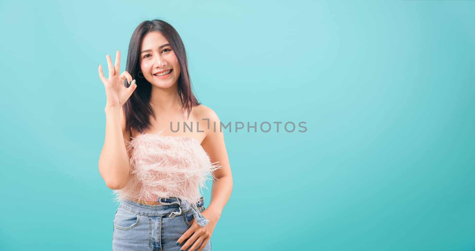 Asian happy portrait beautiful young woman standing smiling showing hand okay sign and looking to camera isolated on blue background with copy space for text