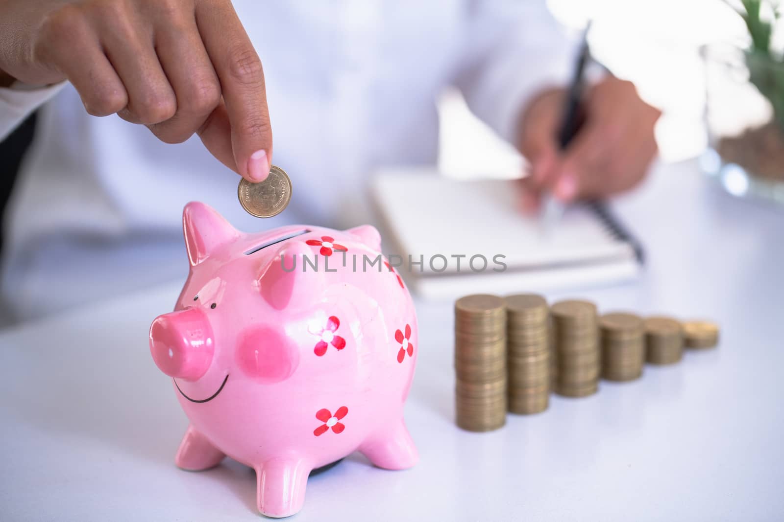 The hand of a business person is going to put a coin in a pink piggy bank. Hand was about to write an income and expense account.Financial accounting concepts. Calculate assets for business success. by Kanjana