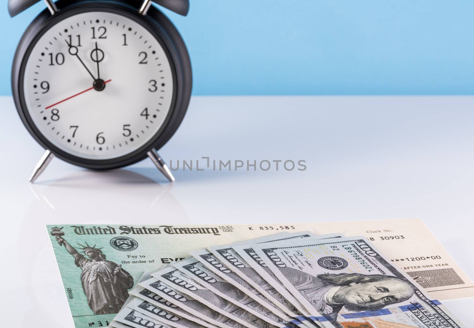 Stack of 100 dollar bills with illustrative coronavirus stimulus payment check and alarm clock to show delay in receiving money