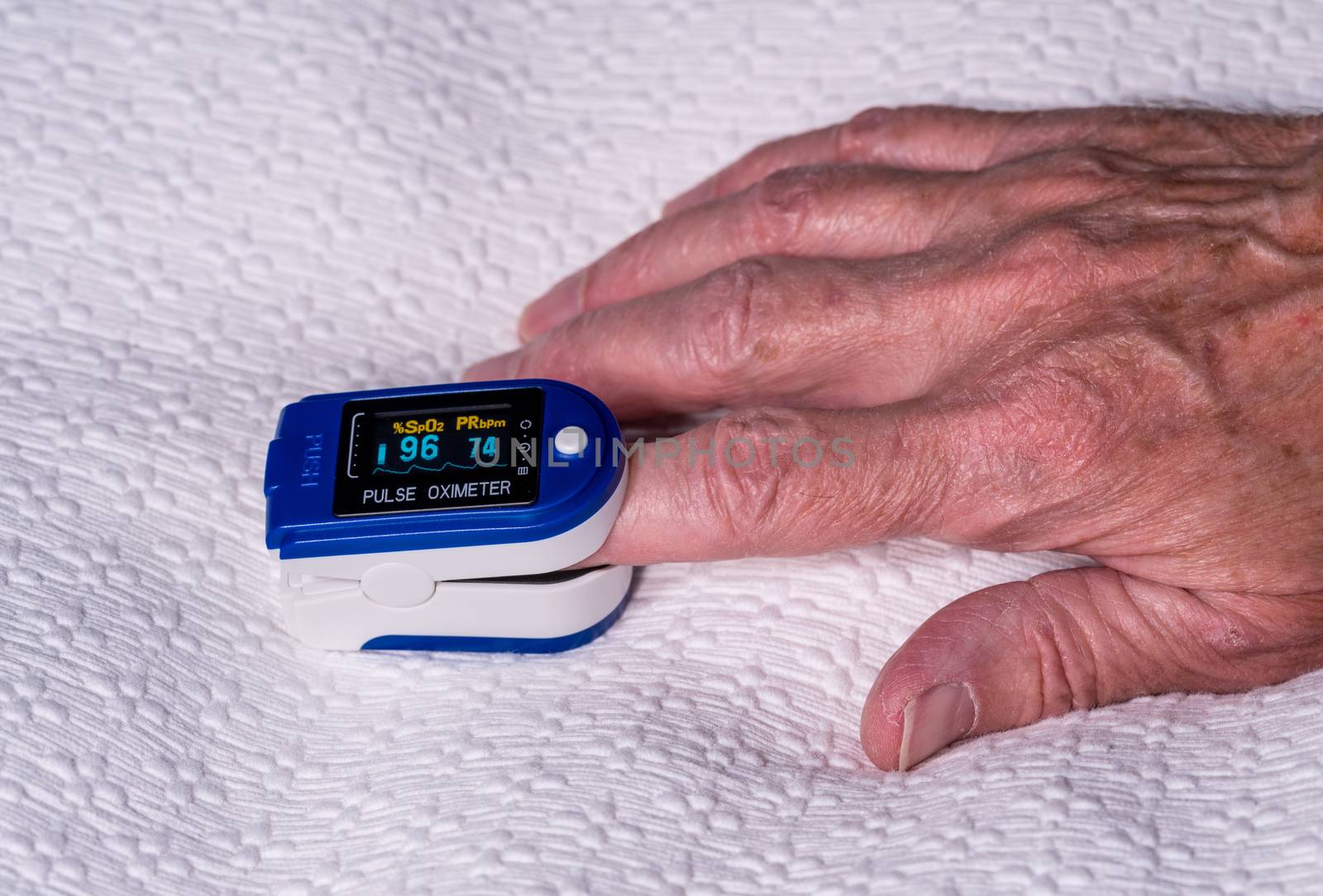 Pulse oximeter on finger used to test blood oxygen level in case of virus infection of the lungs with senior hand resting on bedspread