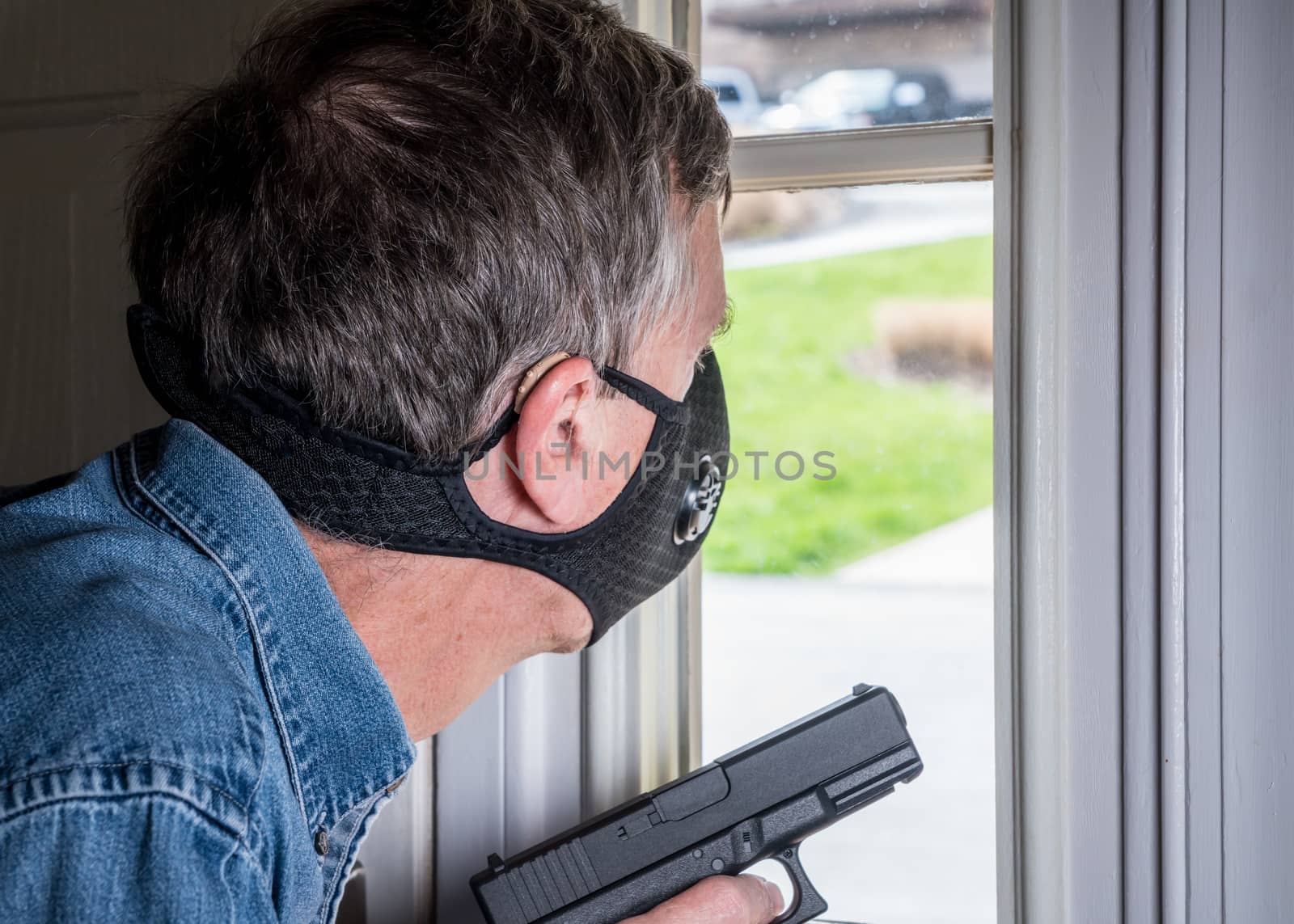 Senior man with face mask looking worried at front door and ready to defend home with a gun during coronavirus quarantine