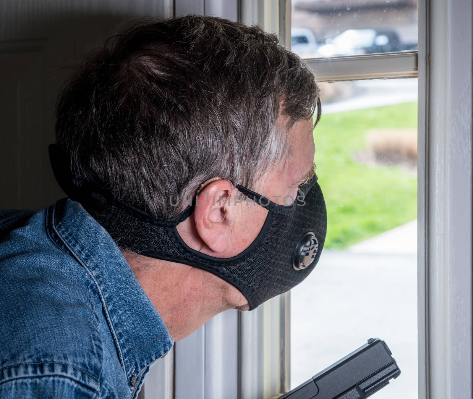 Senior man with face mask looking worried at front door and ready to defend home with a gun during coronavirus quarantine