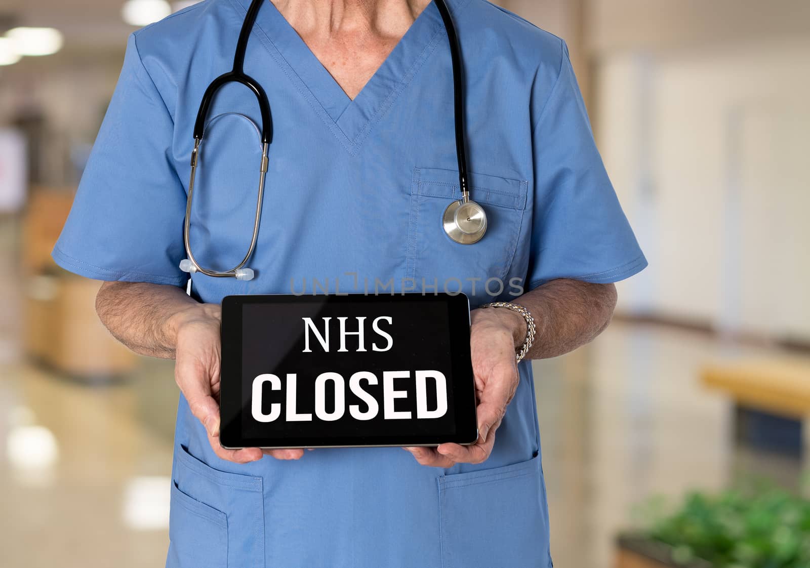 Senior male caucasian doctor with stethoscope in medical scrubs holding electronic tablet for saying NHS hospital is closed due to coronavirus