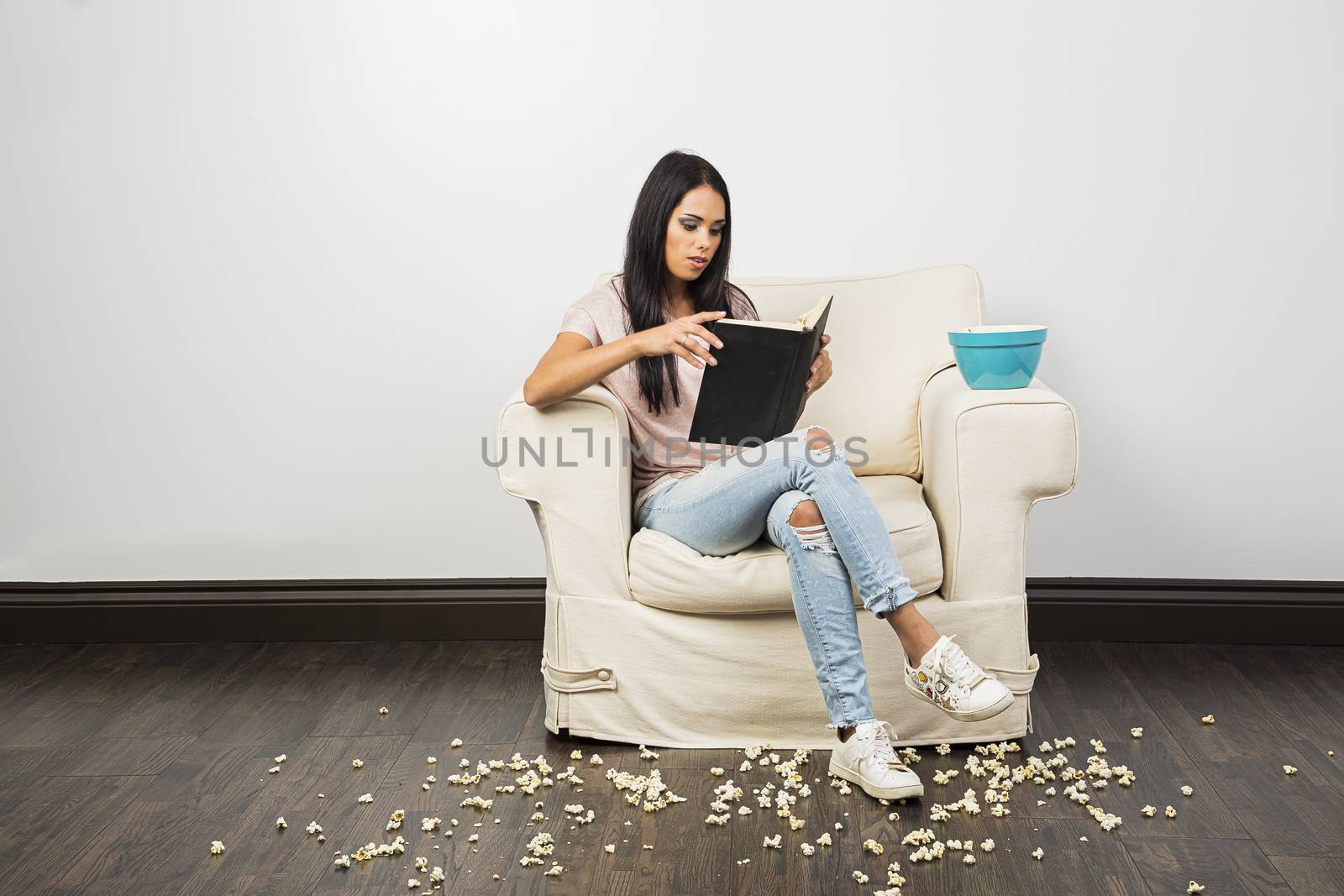 young woman sitting on a couch, reading a hardcover book and eating popcorn while making a mess
