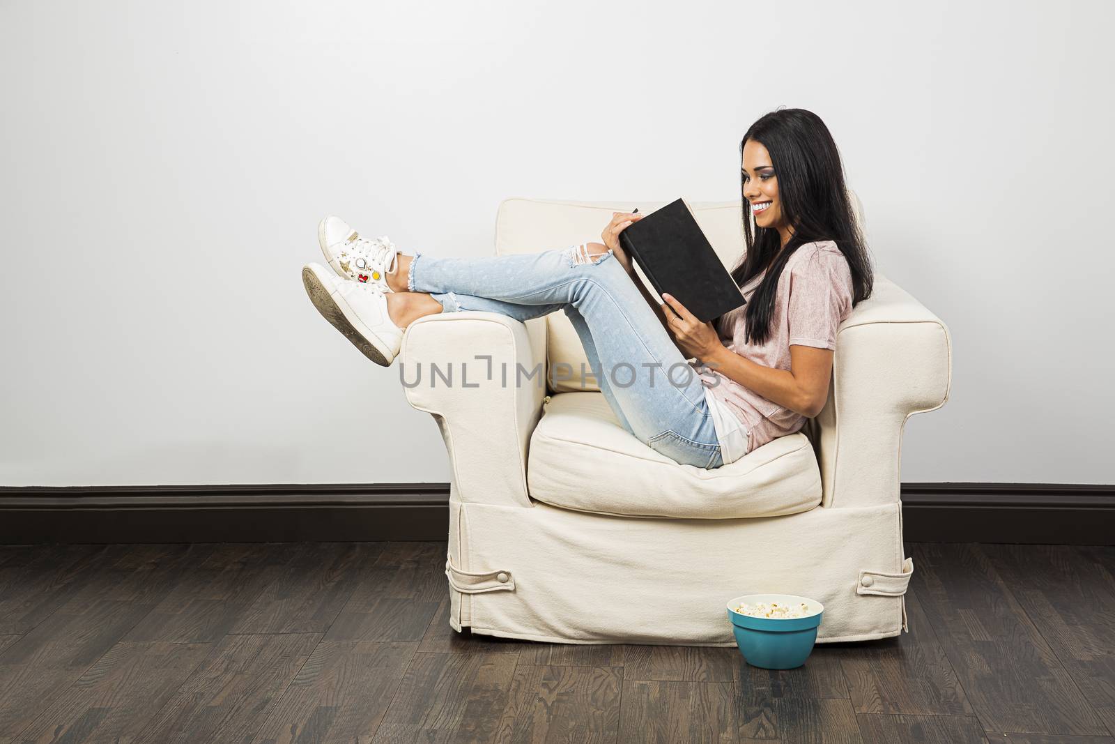 young woman sitting on a couch with her legs up, reading a hardcover book and eating popcorn 

