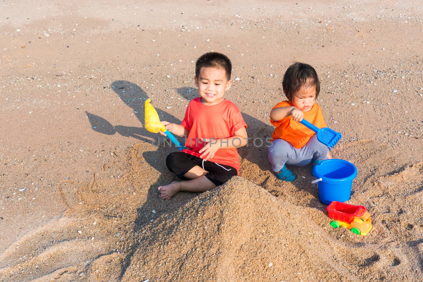 Asian Thai happy cute little cheerful Brother and sister two children funny digging play toy with sand at an outdoor tropical beach in summer day with copy space