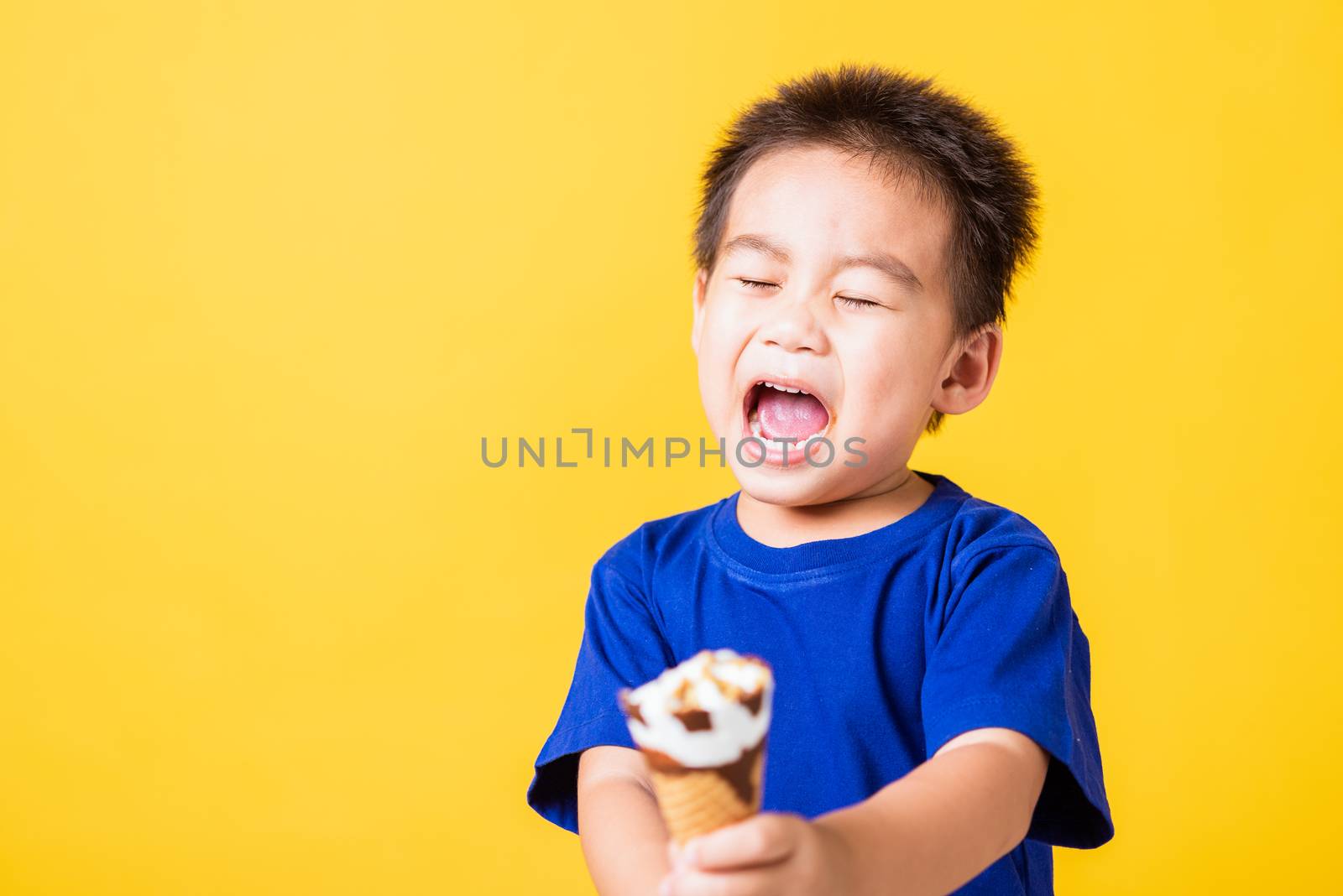 Happy portrait Asian child or kid cute little boy attractive laugh smile playing holds and eating sweet chocolate ice cream waffle cone, studio shot isolated on yellow background, summer concept